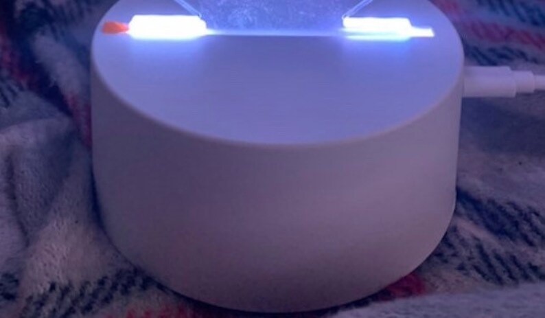 May include: A white, round, charging base with two bright white LED lights on the top. The base is plugged into a white cord.