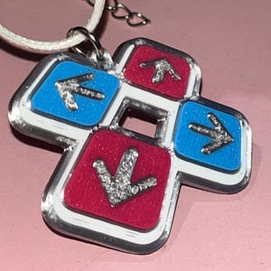 May include: A pendant necklace featuring a dance pad design. The pendant has a white border with red and blue squares, each with a silver arrow. The necklace has a white cord and silver chain.