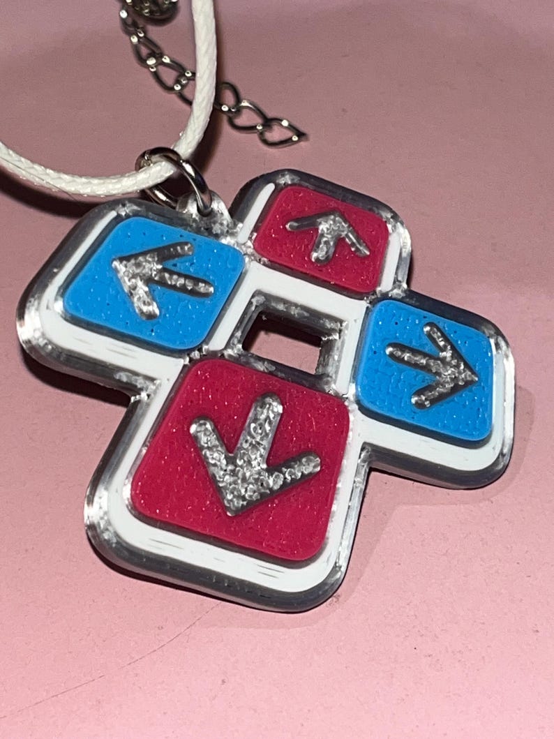 May include: A pendant necklace featuring a dance pad design. The pendant has a white border with red and blue squares, each with a silver arrow. The necklace has a white cord and silver chain.