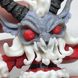 May include: A detailed 3D-printed figurine of a Krampus-like creature. The figure has a gray face with red eyes, white beard and hair, and black horns. It wears a red and white outfit, resembling a Santa Claus costume. The figurine is held in a hand.