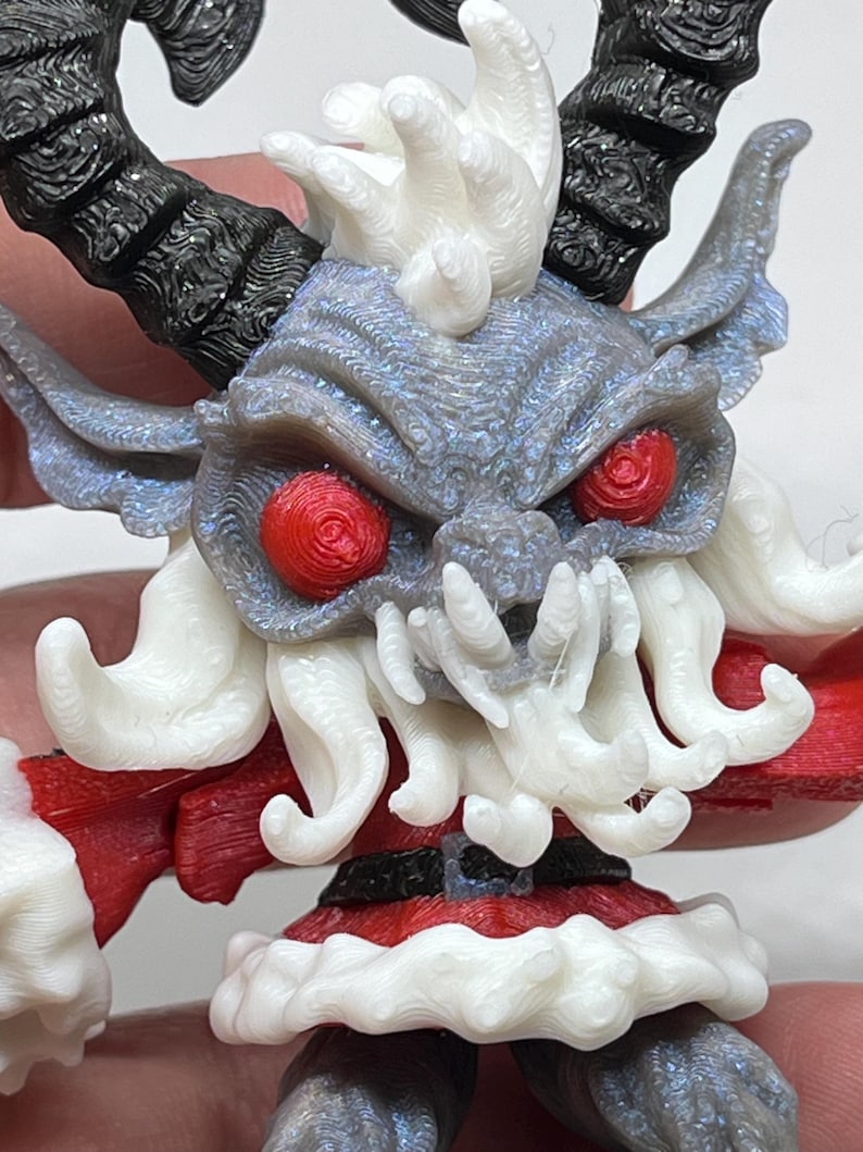 May include: A detailed 3D-printed figurine of a Krampus-like creature. The figure has a gray face with red eyes, white beard and hair, and black horns. It wears a red and white outfit, resembling a Santa Claus costume. The figurine is held in a hand.