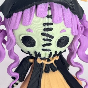 May include: A 3D printed doll with a stitched face, purple hair, and a black witch's hat and cape. The doll is wearing a yellow dress and black and white striped stockings.