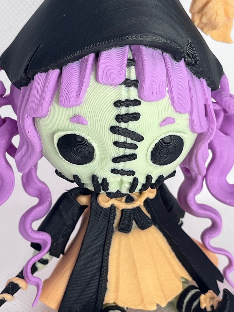 May include: A 3D printed doll with a stitched face, purple hair, and a black witch's hat and cape. The doll is wearing a yellow dress and black and white striped stockings.
