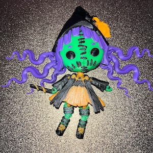 May include: A small, green witch doll with purple hair, a black hat, and a black and orange dress. The doll is holding a stick in its hand.