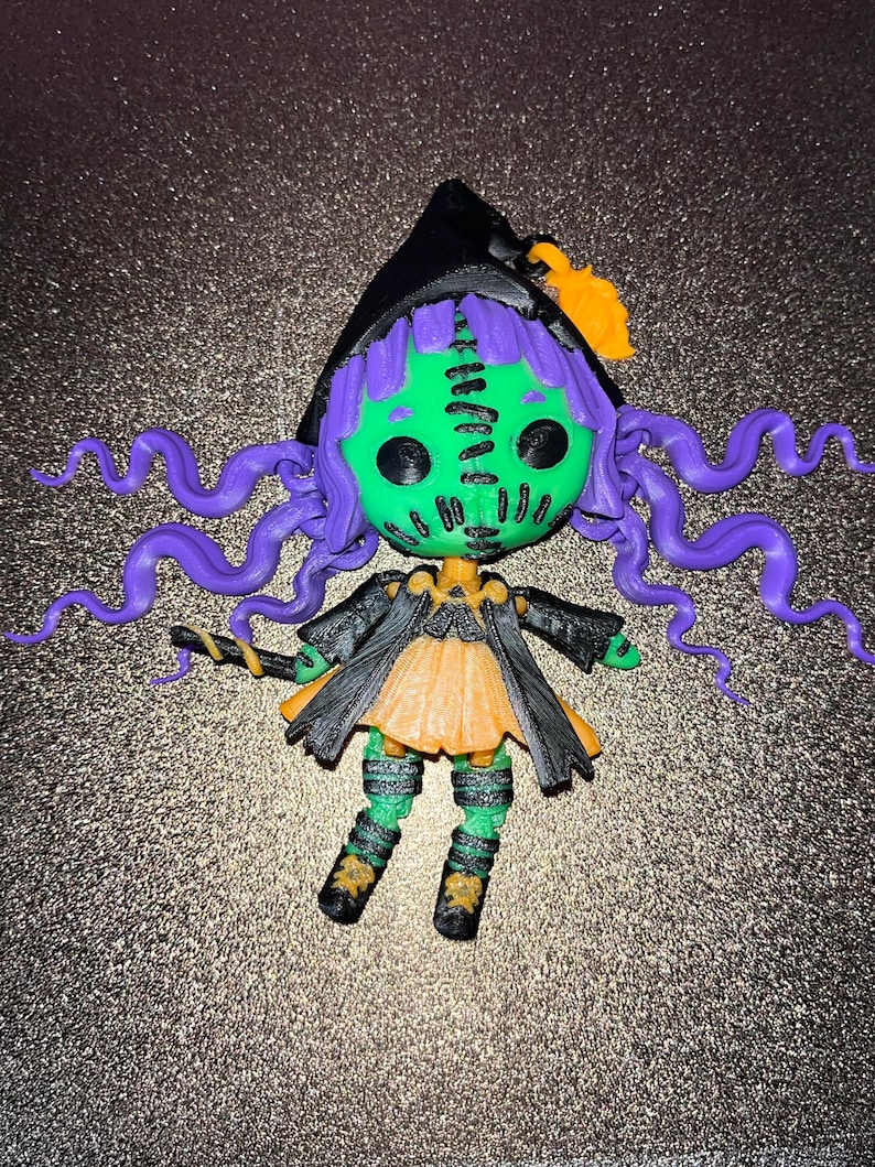 May include: A small, green witch doll with purple hair, a black hat, and a black and orange dress. The doll is holding a stick in its hand.