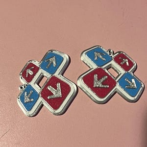 May include: Two silver-edged charms, each featuring a red and blue square design with arrows. The red squares have a glittery texture, and the blue squares have a smooth finish. The charms are set against a pink background.