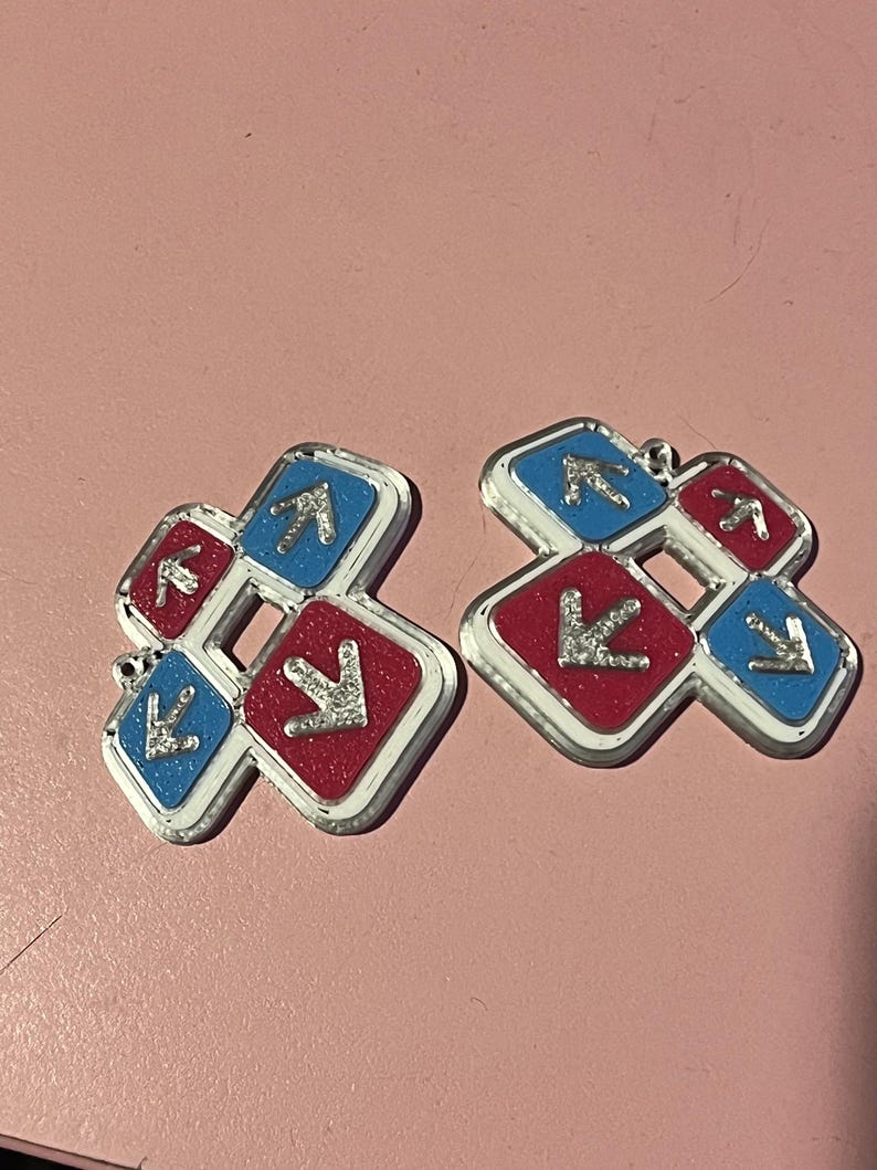 May include: Two silver-edged charms, each featuring a red and blue square design with arrows. The red squares have a glittery texture, and the blue squares have a smooth finish. The charms are set against a pink background.