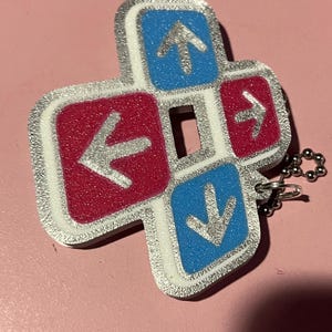 May include: A silver-edged keychain charm in a cross shape, featuring four directional buttons. The buttons are red and blue with silver arrows. A small silver chain is attached.
