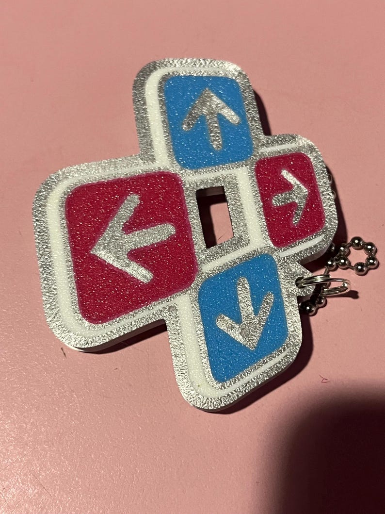 May include: A silver-edged keychain charm in a cross shape, featuring four directional buttons. The buttons are red and blue with silver arrows. A small silver chain is attached.