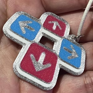 May include: A Dance Dance Revolution-themed pendant with a silver glitter border. The pendant features four directional arrows in blue and red, with a white cord for wearing. The arrows point up, down, left, and right.