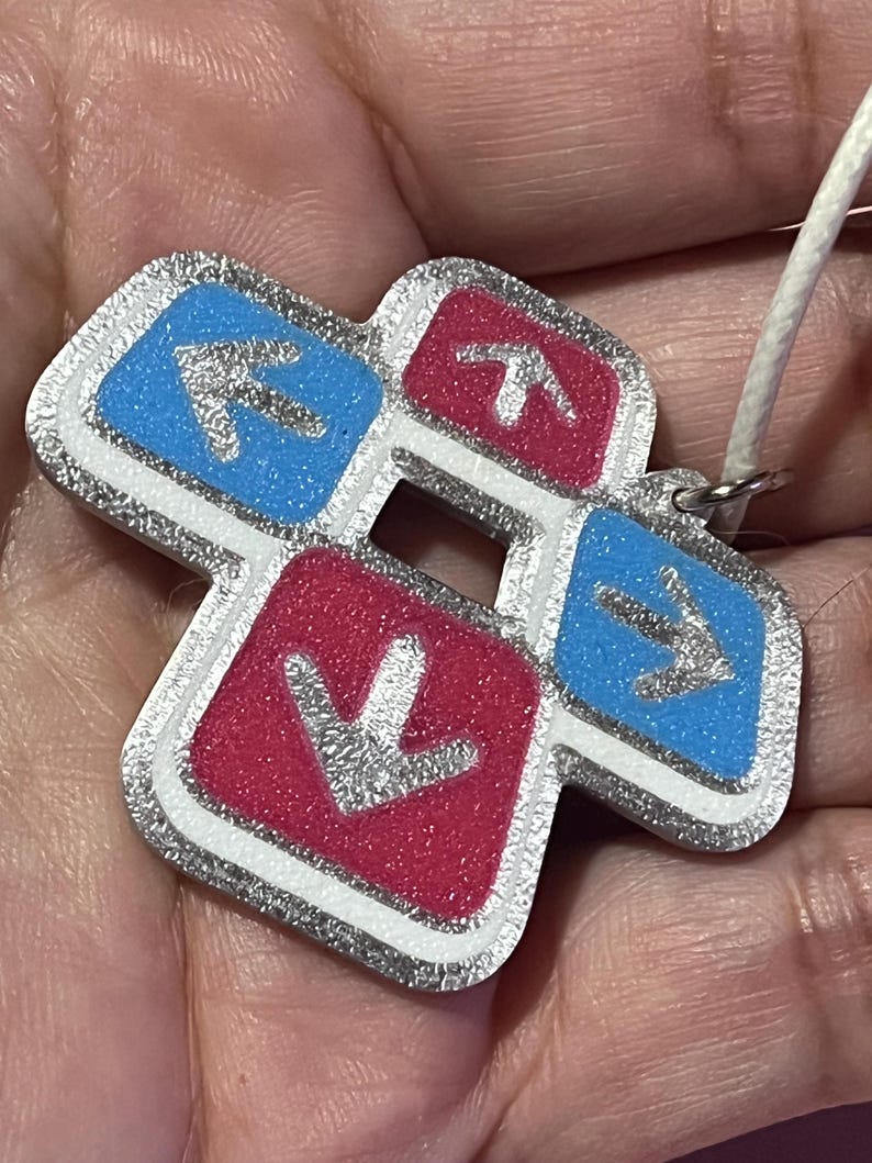 May include: A Dance Dance Revolution-themed pendant with a silver glitter border. The pendant features four directional arrows in blue and red, with a white cord for wearing. The arrows point up, down, left, and right.