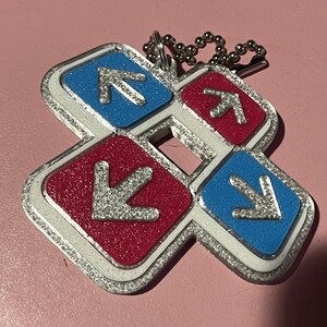 May include: A keychain featuring a dance pad design with four directional arrows. The arrows are silver and set against red and blue backgrounds, outlined in silver. A silver ball chain is attached.
