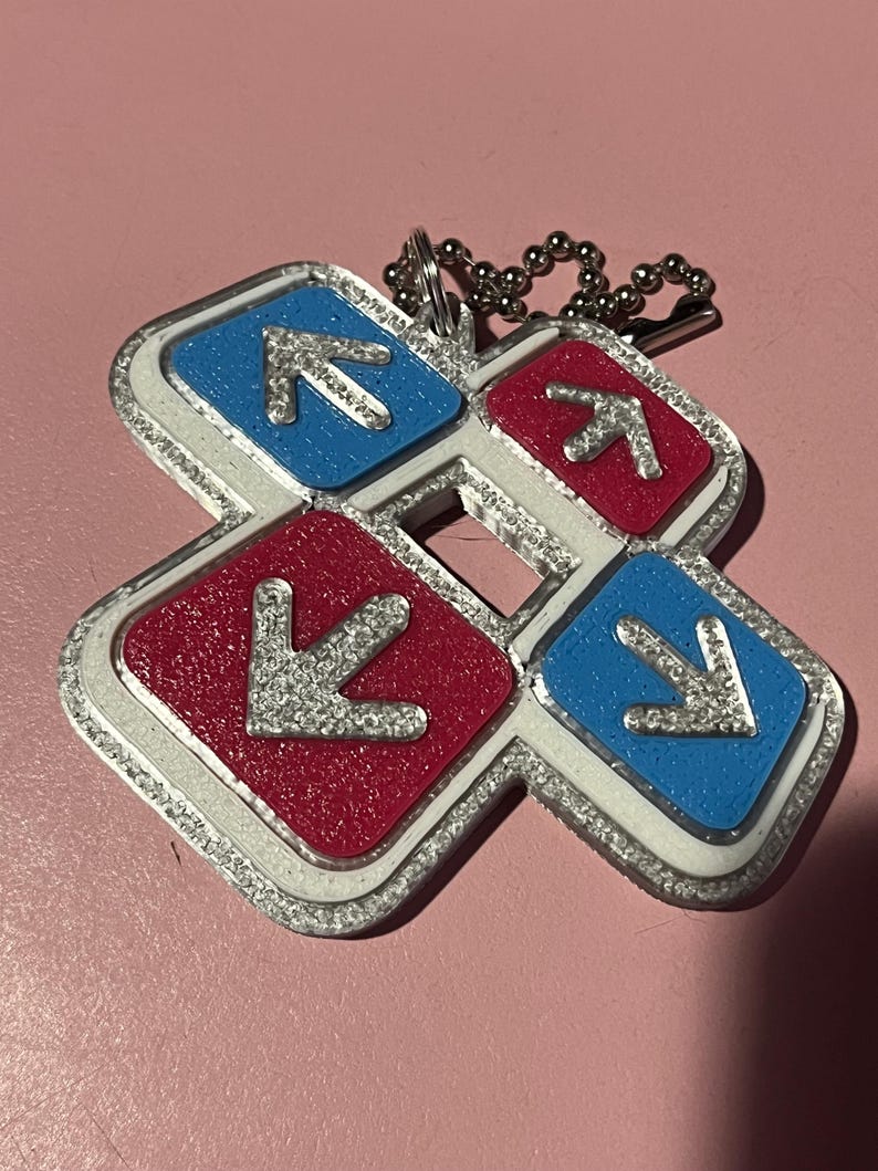 May include: A keychain featuring a dance pad design with four directional arrows. The arrows are silver and set against red and blue backgrounds, outlined in silver. A silver ball chain is attached.