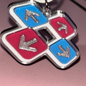 May include: A pendant featuring a dance pad design with red and blue squares, each with a silver arrow. The pendant has a white border and a silver chain. The background is a light pink color.