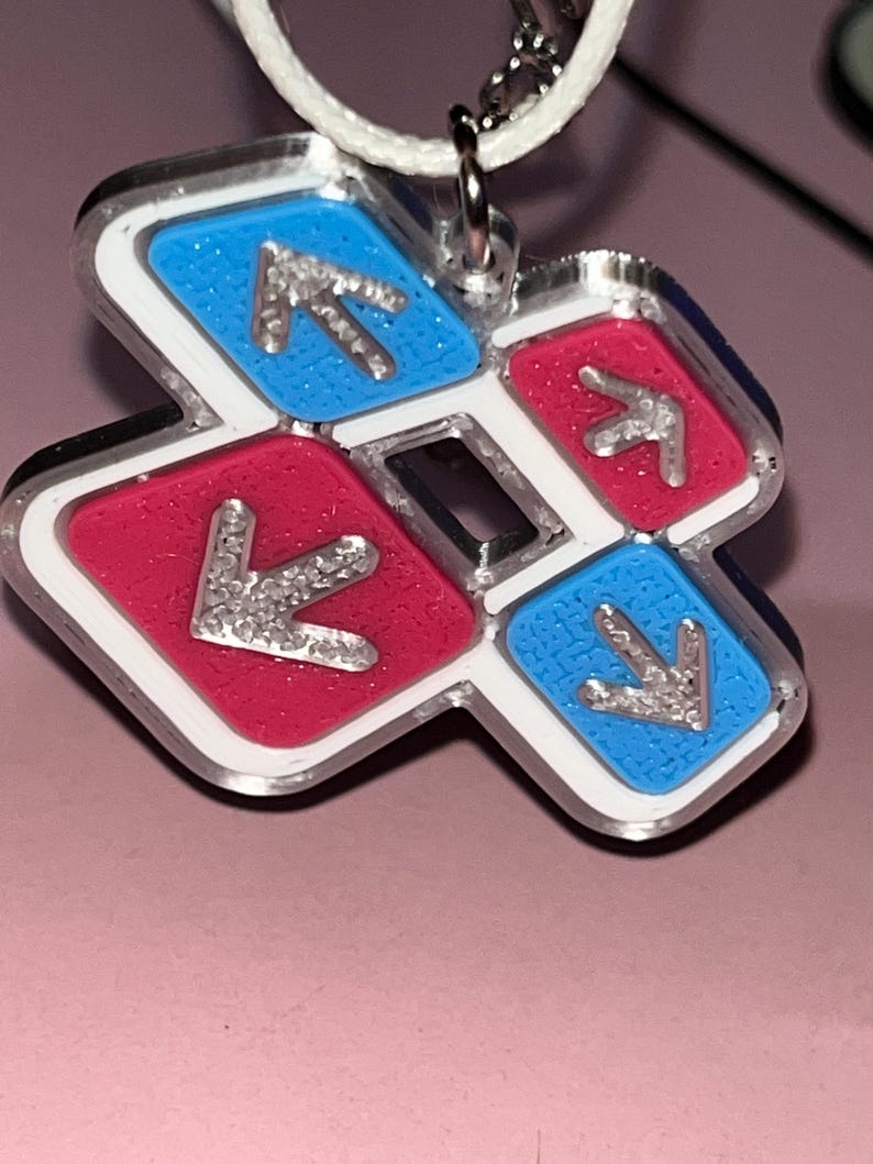 May include: A pendant featuring a dance pad design with red and blue squares, each with a silver arrow. The pendant has a white border and a silver chain. The background is a light pink color.