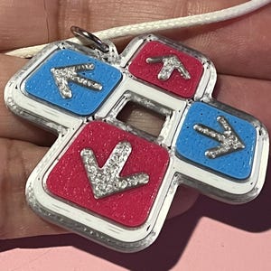 May include: A pendant necklace featuring a dance pad design. The pendant has a silver border with four colored squares: two blue and two red. Each square has a silver arrow. The pendant hangs from a white cord.