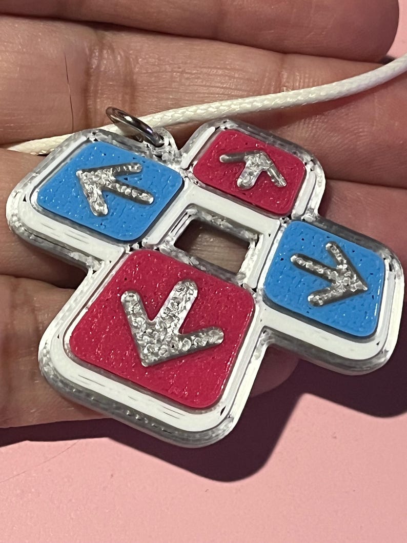 May include: A pendant necklace featuring a dance pad design. The pendant has a silver border with four colored squares: two blue and two red. Each square has a silver arrow. The pendant hangs from a white cord.