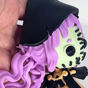 May include: A 3D printed figurine of a witch with long purple hair, wearing a black hat and a yellow dress. The witch has a stitched-up face with a black eye.