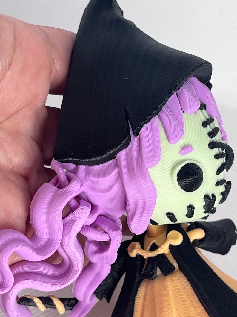May include: A 3D printed figurine of a witch with long purple hair, wearing a black hat and a yellow dress. The witch has a stitched-up face with a black eye.