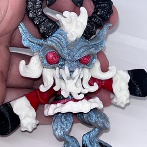 May include: A detailed figurine of a horned creature with red eyes, black horns, and a blue and white body. The figure has a red and white outfit with black hooves and hands. The figurine is held in a hand.
