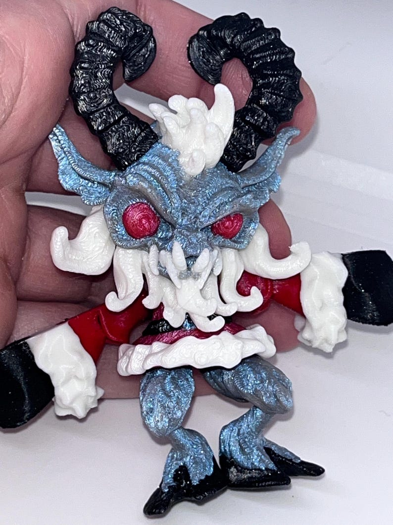 May include: A detailed figurine of a horned creature with red eyes, black horns, and a blue and white body. The figure has a red and white outfit with black hooves and hands. The figurine is held in a hand.
