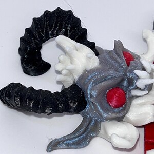 May include: A detailed, 3D-printed sculpture of a horned creature's head. The horns are black, while the head features gray, white, and red accents. The eyes are a vibrant red, and the overall design has a fantasy theme.