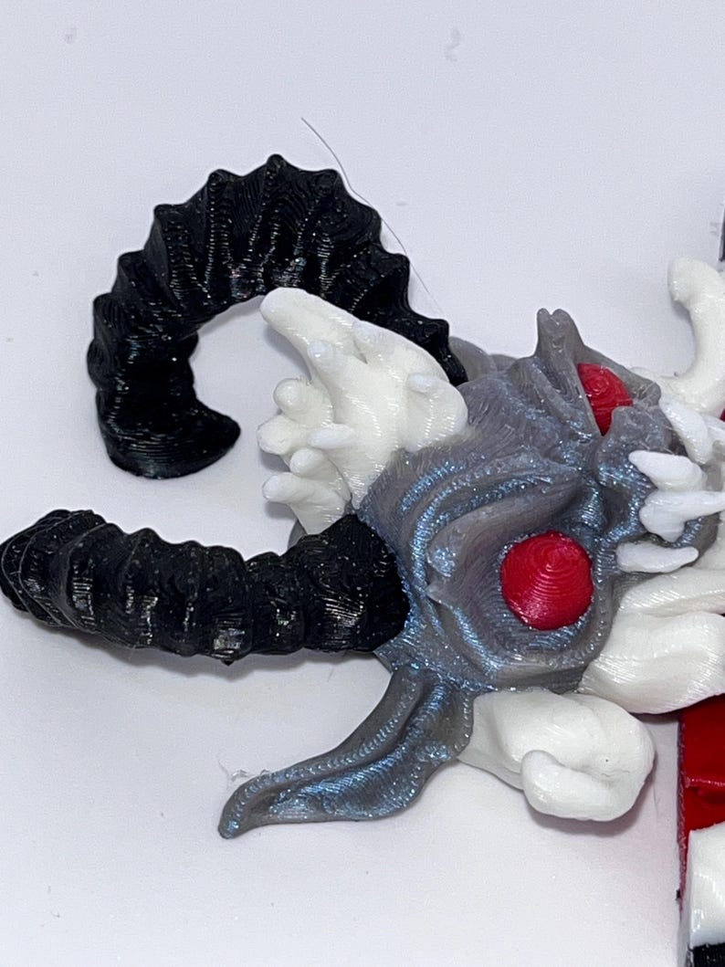 May include: A detailed, 3D-printed sculpture of a horned creature's head. The horns are black, while the head features gray, white, and red accents. The eyes are a vibrant red, and the overall design has a fantasy theme.
