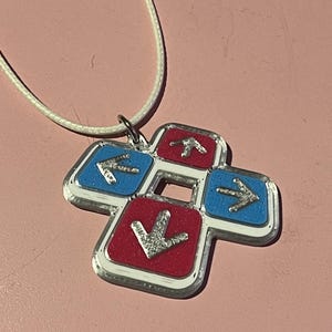 May include: A pendant necklace featuring a four-arrow design in red and blue, with silver arrows and a white cord. The pendant has a silver border and is likely made of metal or a similar material. The arrows point up, down, left, and right.