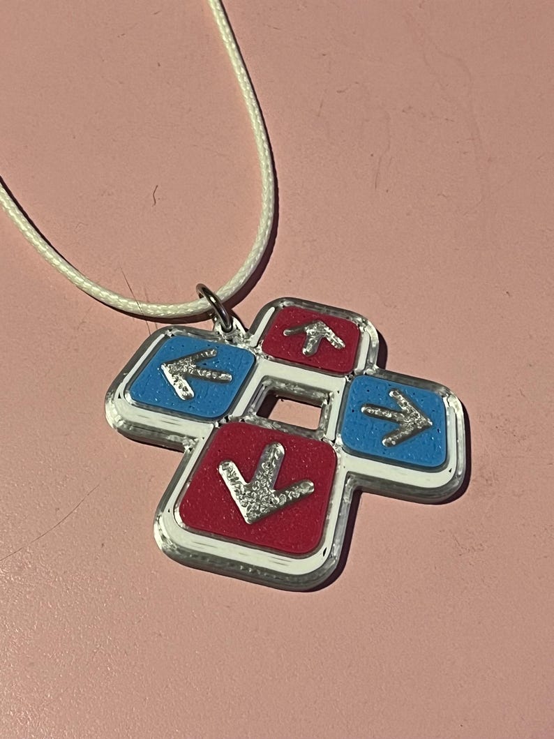 May include: A pendant necklace featuring a four-arrow design in red and blue, with silver arrows and a white cord. The pendant has a silver border and is likely made of metal or a similar material. The arrows point up, down, left, and right.