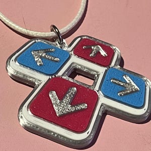May include: A pendant necklace featuring a four-arrow design. The pendant has a white border with red and blue squares, each with a silver arrow. The necklace has a white cord and a silver clasp. The background is a light pink.