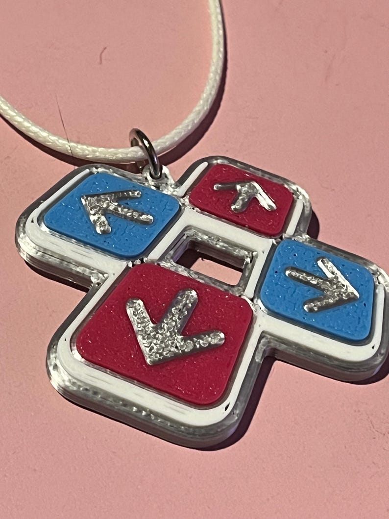 May include: A pendant necklace featuring a four-arrow design. The pendant has a white border with red and blue squares, each with a silver arrow. The necklace has a white cord and a silver clasp. The background is a light pink.