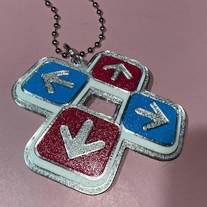 May include: A silver-colored keychain featuring a dance pad design. The pad has four directional arrows in red and blue, with a silver border and a ball chain for attachment. The arrows point up, down, left, and right.