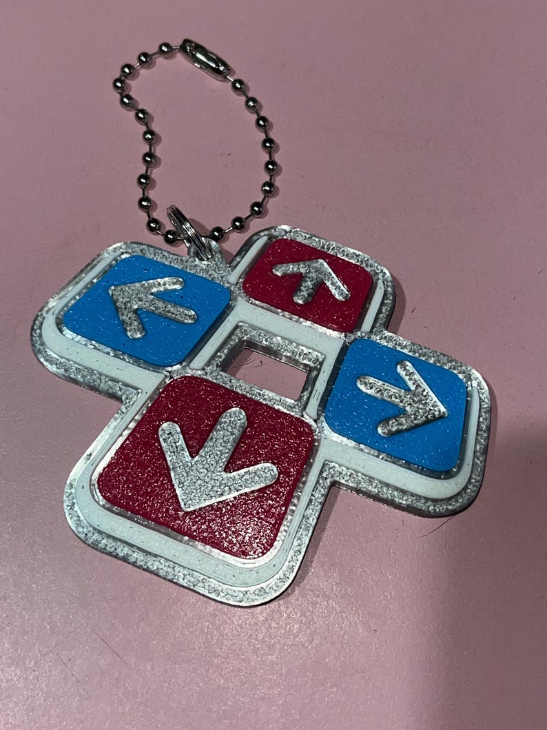 May include: A silver-colored keychain featuring a dance pad design. The pad has four directional arrows in red and blue, with a silver border and a ball chain for attachment. The arrows point up, down, left, and right.