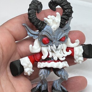 May include: A detailed figurine of a Krampus-like creature. The figure has black horns, red eyes, and a white beard and trim. The creature is wearing a red outfit with white trim and black gloves and boots. The figure is held in a hand.