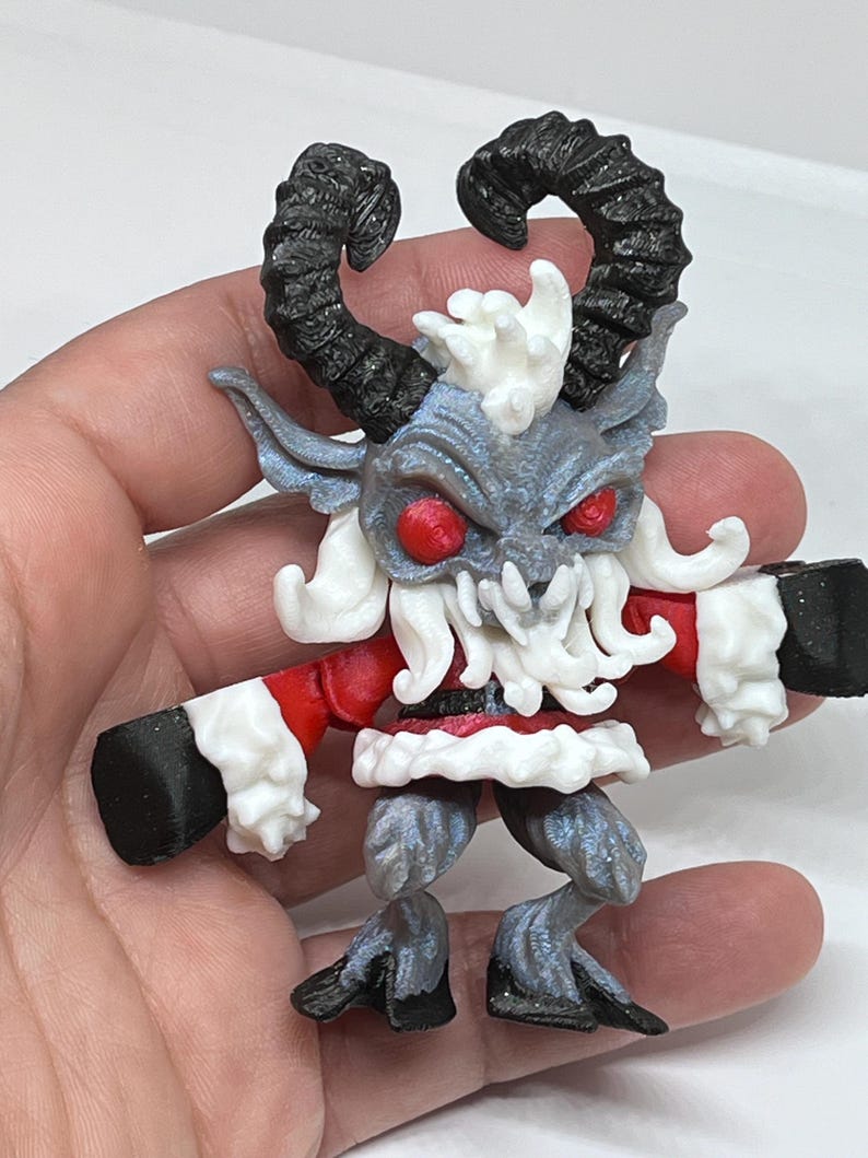 May include: A detailed figurine of a Krampus-like creature. The figure has black horns, red eyes, and a white beard and trim. The creature is wearing a red outfit with white trim and black gloves and boots. The figure is held in a hand.