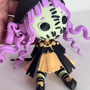 May include: A small, 3D printed doll with a stitched face, purple hair, a black cape, and a yellow dress. The doll is wearing black and white striped stockings and black shoes.