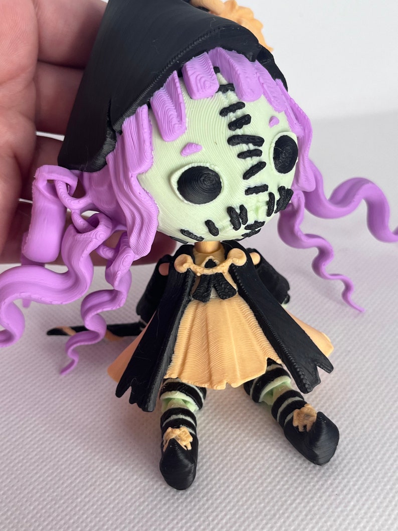 May include: A small, 3D printed doll with a stitched face, purple hair, a black cape, and a yellow dress. The doll is wearing black and white striped stockings and black shoes.