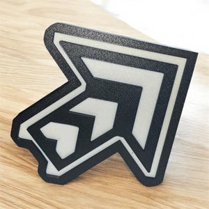 May include: A black and white arrow-shaped object with a textured surface. The arrow has a layered design with a white interior and a black outline. The object is on a light wood surface.