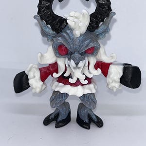 May include: A detailed figurine of a Krampus-like creature. It features large, curved black horns, red eyes, and a menacing expression. The figure is primarily gray with white and red accents, including a red coat with white trim. The feet and hands are black.