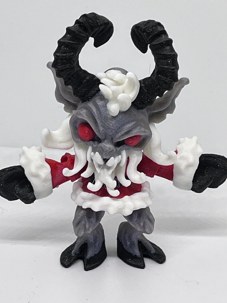 May include: A 3D-printed figurine of a Krampus-like creature. It has black horns, red eyes, and a gray face. The figure wears a red coat with white trim and black hooves. The creature has a menacing expression.