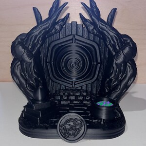 May include: A black 3D printed model of a portal with a cauldron and a circular emblem. The portal is surrounded by a black, twisted, tree-like structure. The cauldron is filled with green and purple liquid. The emblem has the words "Inkling Press" on it.