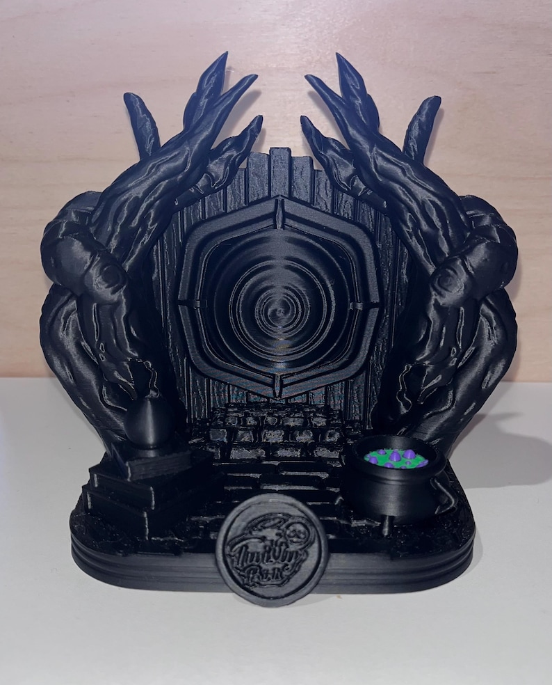 May include: A black 3D printed model of a portal with a cauldron and a circular emblem. The portal is surrounded by a black, twisted, tree-like structure. The cauldron is filled with green and purple liquid. The emblem has the words "Inkling Press" on it.