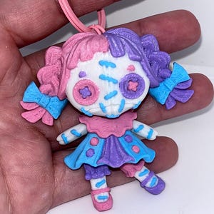 May include: A pink and purple doll keychain with a white face, blue eyes, and a pink and blue dress. The doll has a stitched mouth and a blue bow on each side of its head.