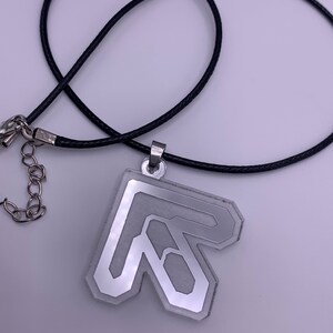 Puede incluir: Un collar con un colgante de plata con un diseño de letra R estilizada. El colgante está unido a un cordón negro.