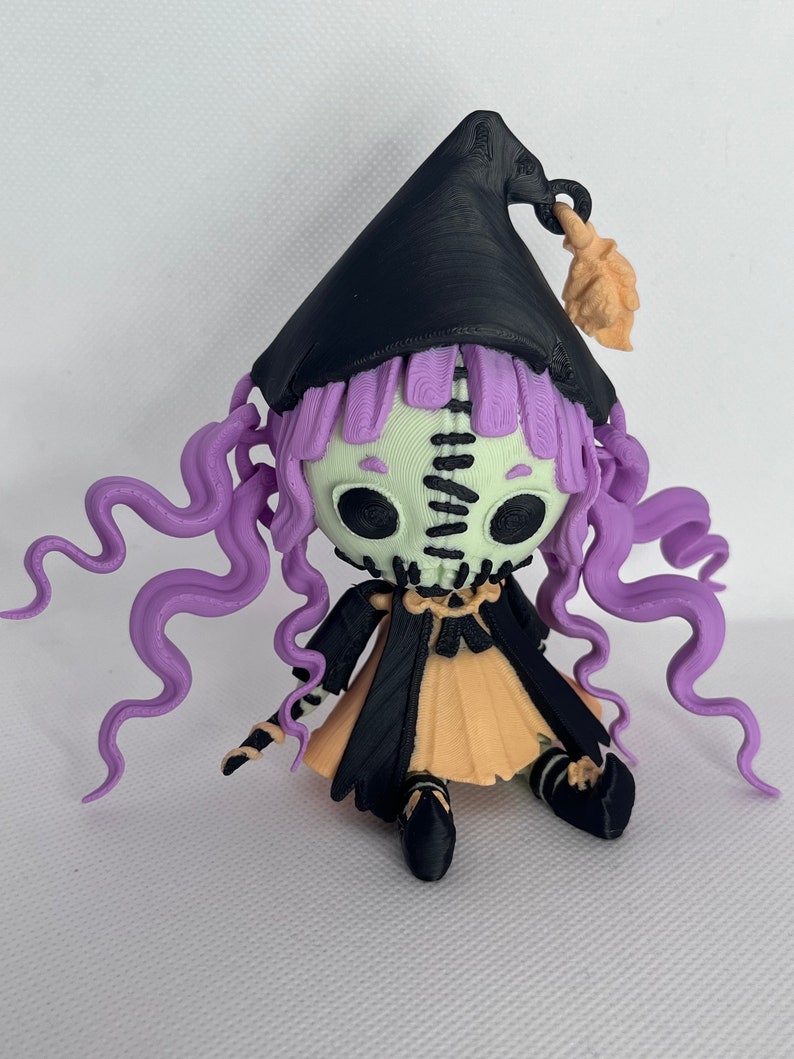 May include: A 3D printed figurine of a witch with a black pointed hat, purple hair, and a green and black skeleton face. The witch is wearing a black and orange dress and black shoes.