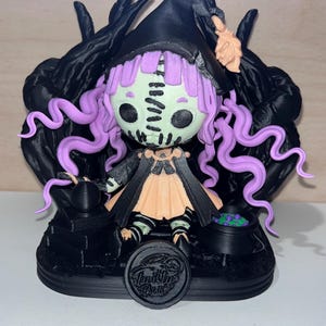 May include: A 3D printed figurine of a witch with purple hair, wearing a black hat and a peach-coloured dress. The witch is sitting on a black base with a cauldron and a small cauldron with purple liquid. The base has a black circle with the text "Ink the Park" in white.