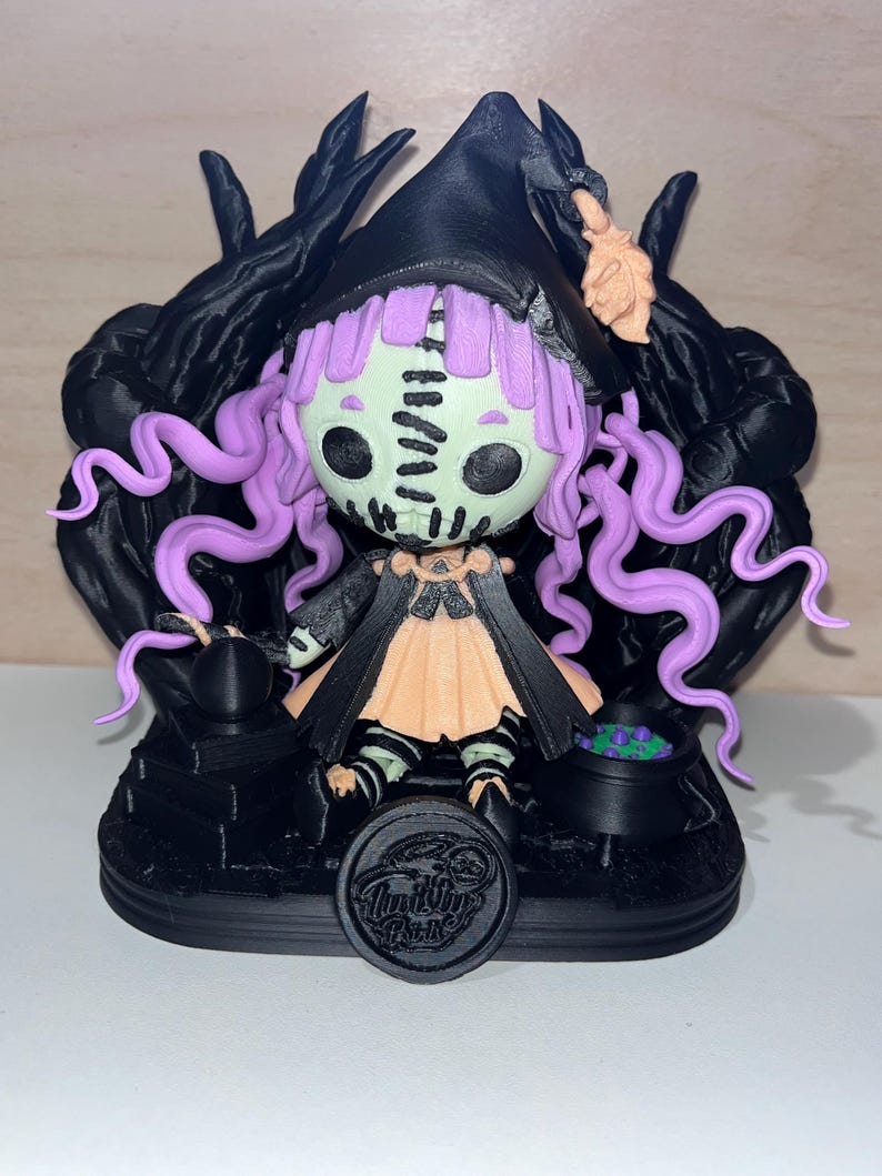 May include: A 3D printed figurine of a witch with purple hair, wearing a black hat and a peach-coloured dress. The witch is sitting on a black base with a cauldron and a small cauldron with purple liquid. The base has a black circle with the text "Ink the Park" in white.