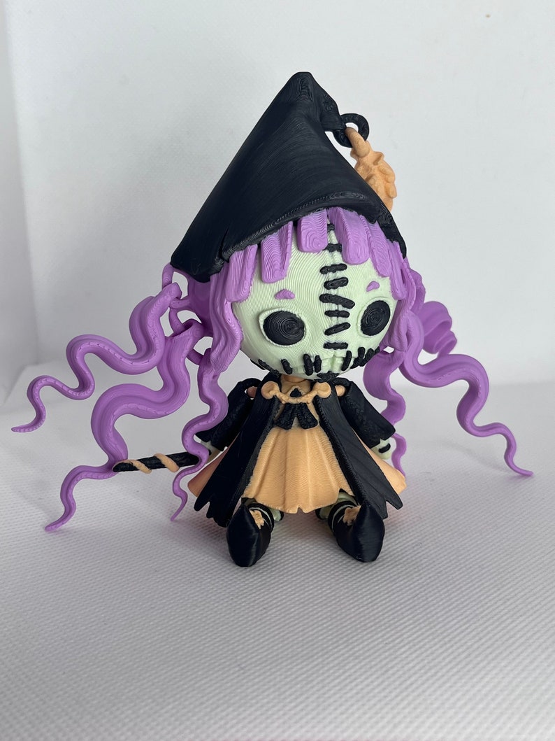 May include: A 3D printed figurine of a witch with purple hair, a black hat, and a black and orange dress. The witch is holding a stick in her hand.