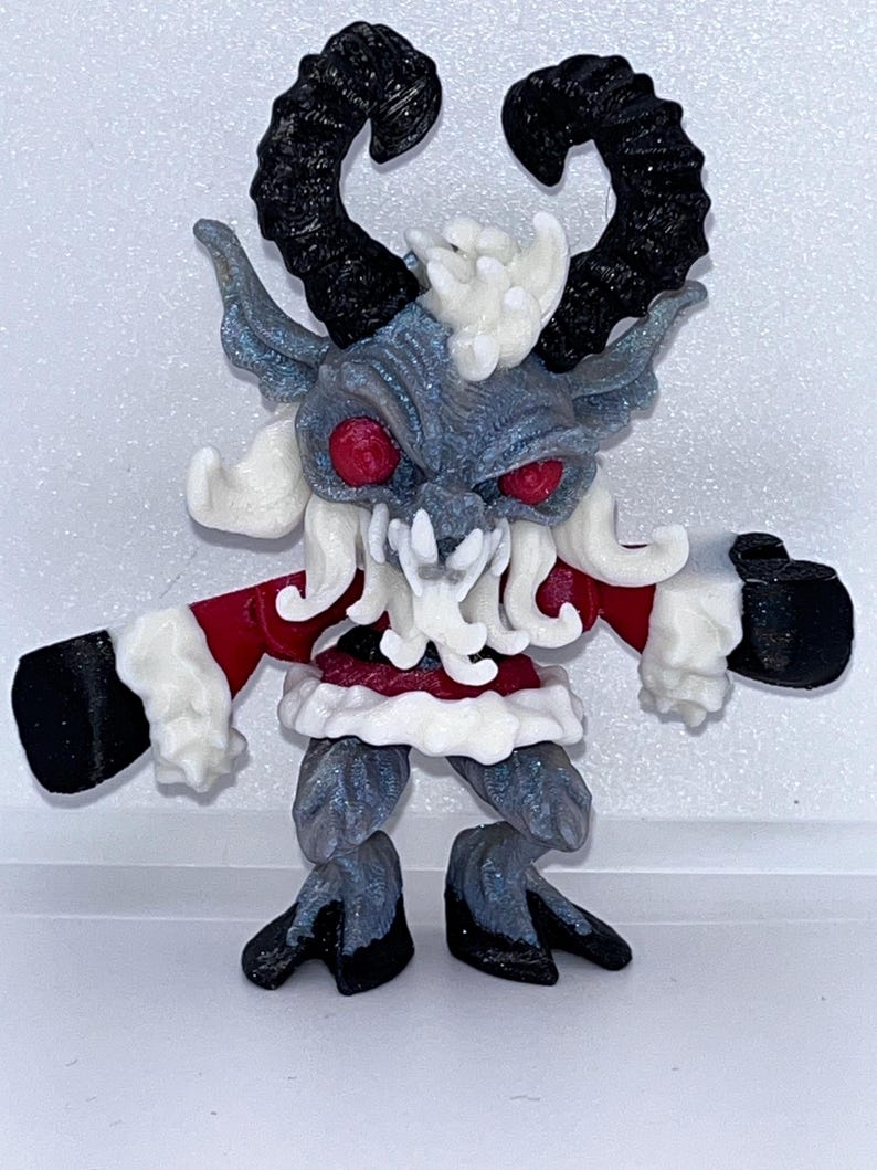 May include: A figurine of a Krampus-like creature with black horns, red eyes, and a red and white outfit. The figure has a gray body with white accents and black hooves and gloves. The figurine is standing upright.