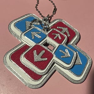 May include: A keychain featuring a layered design of red and blue squares with silver arrows and a silver border. The keychain has a silver chain for attachment. The background is a solid pink color.